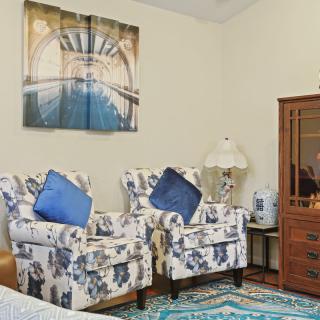 A cozy living room featuring two patterned armchairs with blue pillows, a scenic artwork on the wall, a colorful rug, and a wooden cabinet next to a decorative lamp.