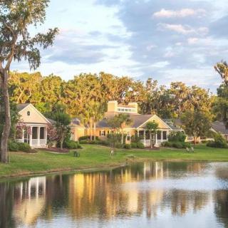 Charming lakeside houses surrounded by trees and reflected in the water under a partly cloudy sky.