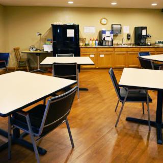 An empty cafeteria with tables and chairs, featuring a small kitchen area in the background.