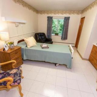 A cozy and simple bedroom featuring a bed, nightstand, armchair, and a window with curtains, providing a peaceful atmosphere.
