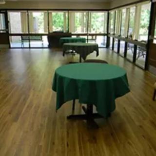 A spacious room with round tables covered in green tablecloths and surrounded by windows allowing natural light.
