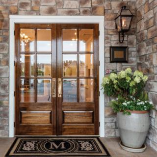 Elegant wooden double doors with a glass pane design, framed by a stone wall with a welcoming doormat and potted flowers.