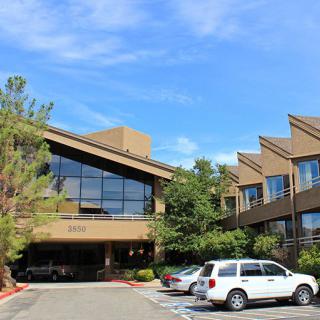 A modern office building with large windows and a parking lot in front, surrounded by trees under a clear blue sky.