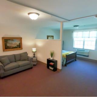 A cozy studio apartment featuring a living area with a sofa and lamp, adjacent to a bedroom space with a bed and window.