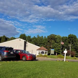 A view of a parking lot with cars in front of a building, surrounded by greenery and under a partly cloudy sky.