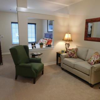 A cozy living room with a green armchair, a beige couch adorned with patterned pillows, and a warm lamp beside a mirror on the wall.