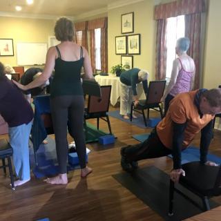A group of adults participates in a yoga class using chairs for support.