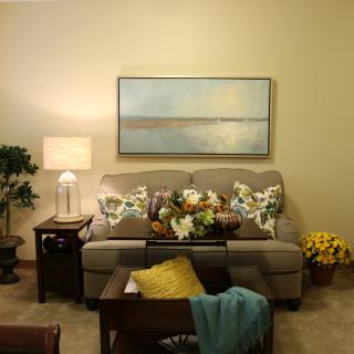 A cozy living room with a sofa adorned with floral cushions, a lamp on a side table, and artwork on the wall.