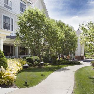 A well-maintained pathway lined with trees and bushes leads to a large residential building on a sunny day.