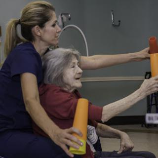 A caregiver assisting an elderly woman with a physical therapy exercise, utilizing colorful foam rollers.