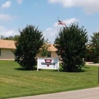 A building with a sign in front, surrounded by trees and a lawn, featuring an American flag against a blue sky.