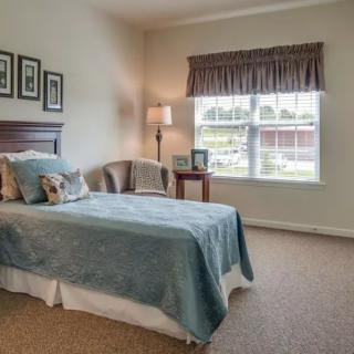 A cozy bedroom with a single bed, carpeted floor, and natural light coming through the window.