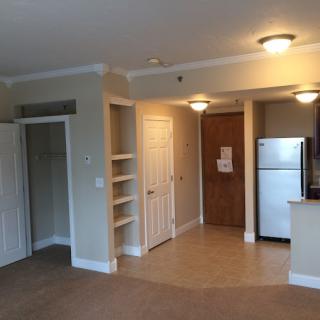 An empty apartment interior featuring a small kitchen with a fridge, cabinets, and light fixtures.