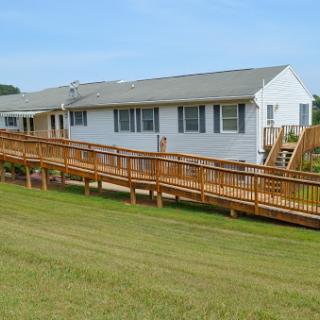 A wooden ramp leads up to a single-story building, surrounded by green grass and under a clear blue sky.