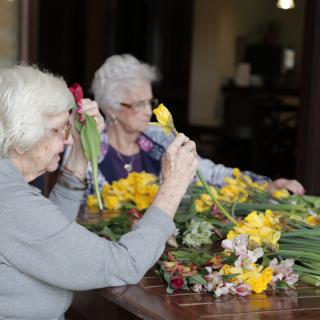 Two elderly women arranging flowers at a table indoors.