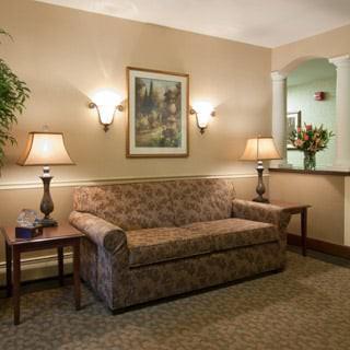 A traditionally decorated living room with a patterned sofa, two lamps on side tables, a framed artwork on the wall, and indoor plants.