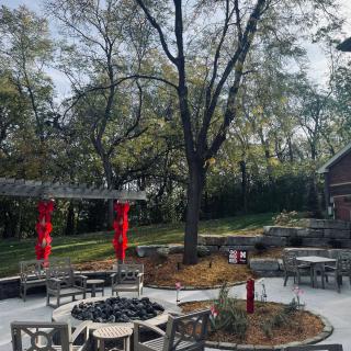 A cozy outdoor patio area featuring a central fire pit, surrounded by trees and equipped with tables and chairs.