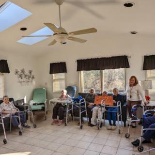 A group of elderly people in a nursing home room, some using wheelchairs and walkers.