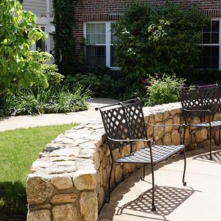 A peaceful garden area featuring metal chairs alongside a curved stone wall, surrounded by lush greenery.