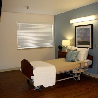 A clean and well-arranged hospital room featuring a single bed with cozy pillows, a lamp on a nightstand, and a window with blinds.