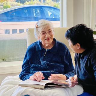 An elderly woman and a young man share a conversation while looking at a book by the window.
