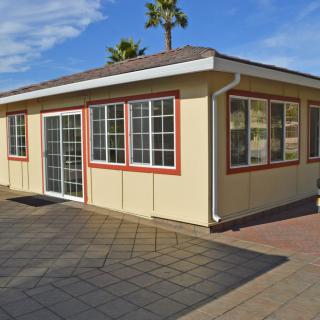 A small building with large windows and a patio area, surrounded by palm trees on a sunny day.