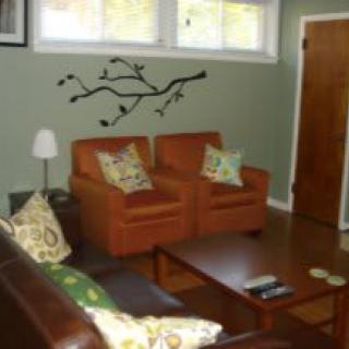 A cozy living room featuring orange chairs, a dark brown sofa, and a wooden coffee table. The walls are adorned with branch-themed wall art.