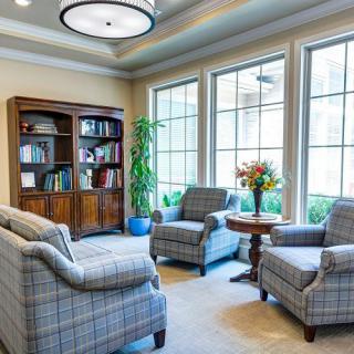 A cozy living room with a bookshelf and four plaid armchairs arranged around a small round table, next to large windows.