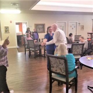 A caregiver interacts with an elderly resident in a nursing home, surrounded by other residents and staff.