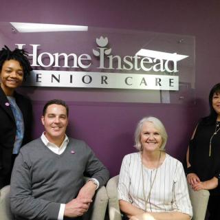 A group of people posing in an office setting with a 'Home Instead Senior Care' sign in the background.
