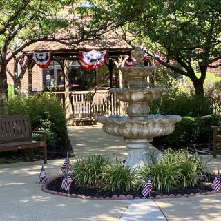 A scenic view of a park with a decorative fountain and gazebo, adorned with patriotic bunting and surrounded by benches.