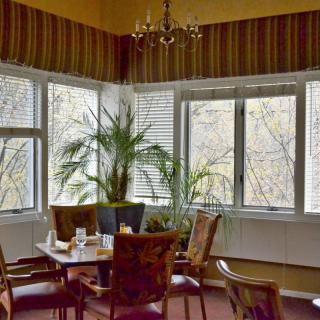 A cozy dining room with a table set for four, surrounded by large windows and lush green plants.