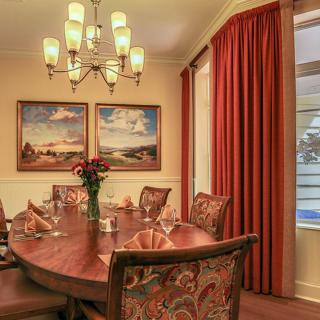 An elegant dining room with a wooden table, surrounded by chairs, and lit by a chandelier. The room features red curtains and landscape paintings on the wall.