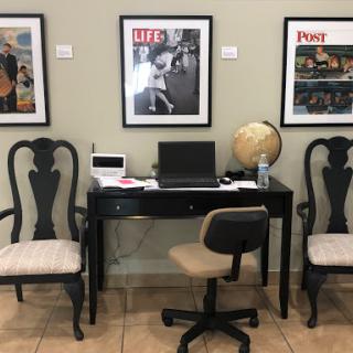 A cozy office space featuring a desk with a computer, two elegant chairs, and framed magazine covers on the wall.