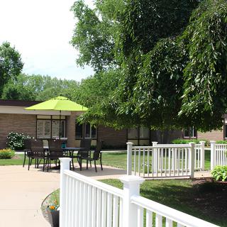 A peaceful garden patio with a green umbrella and benches surrounded by trees and a white fence.
