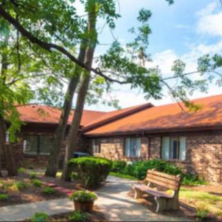 A charming brick house with a red roof surrounded by trees and greenery, featuring a wooden bench in the garden.