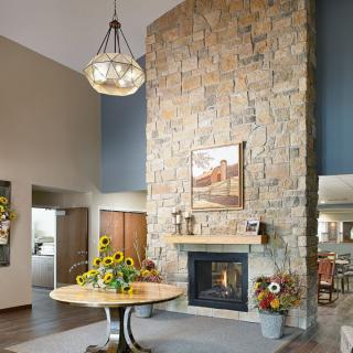 A cozy living room featuring a tall stone fireplace, a chandelier, and a central table with sunflowers.