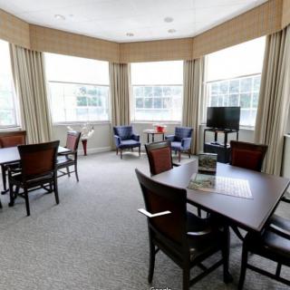 A bright dining room with multiple tables and chairs, featuring large windows with curtains.