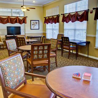A cozy room with tables, chairs, and a TV, featuring large windows with red valances.