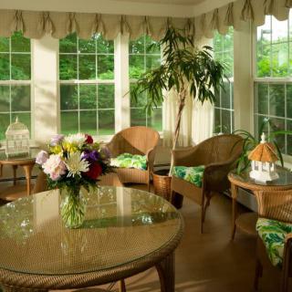 A sunroom featuring wicker furniture and a glass table, adorned with a vibrant flower arrangement, surrounded by large windows and indoor plants.