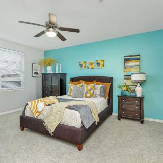 A modern bedroom with a blue accent wall, featuring a bed adorned with decorative pillows, a nightstand with a lamp, and a ceiling fan.