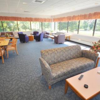 A spacious lounge area with sofas, chairs, and tables, featuring large windows and carpet flooring.
