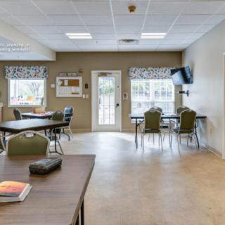 A well-lit community room featuring multiple tables and chairs, with a television mounted on the wall.