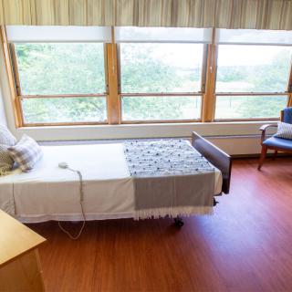 A well-lit hospital room with a single bed and large windows offering a view of greenery.