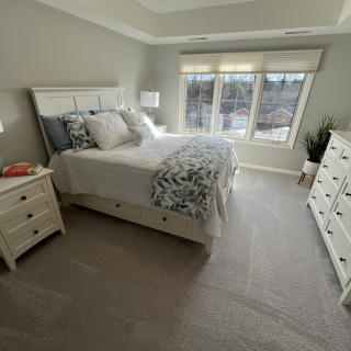 A cozy bedroom with a neatly made bed, two nightstands, and a dresser bathed in natural light from large windows.