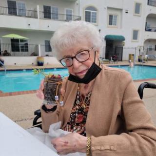 An elderly woman sits by the pool holding a dessert, with a face mask resting below her chin.