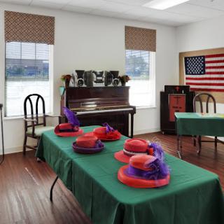 A bright classroom featuring colorful hats on green tables, a piano, and an American flag.