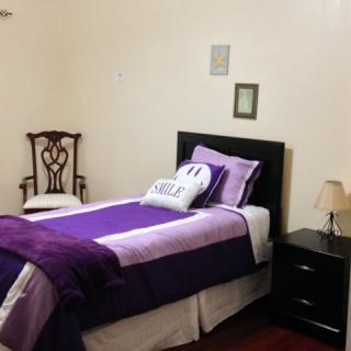A neatly arranged bedroom with a single bed featuring purple bedding, accompanied by a wooden chair and a nightstand with a lamp.