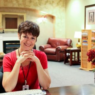A woman in a red shirt is sitting at a desk in an office area, smiling at the camera.