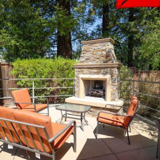 A cozy outdoor patio featuring comfortable seating and a charming stone fireplace, surrounded by lush greenery.